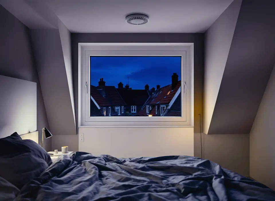 Een slaapkamer in de schemering met een rond ventiel in het plafond en uitzicht op daken en schoorstenen