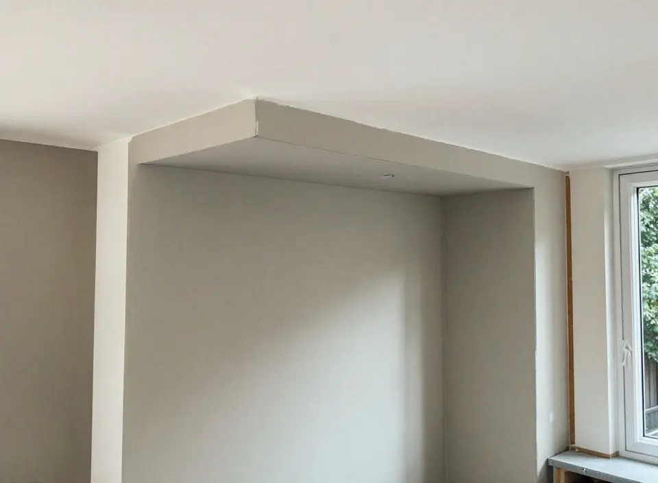 Een nieuwe koof van gipsplaat langs het plafond van een kamer, waarachter de luchtkanalen lopen, met een raam aan de zijkant